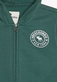Veste zippée verte en tissu doux. Présente un patch logo rond avec "Abercrombie" et un motif d'orignal en blanc. Texture lisse.