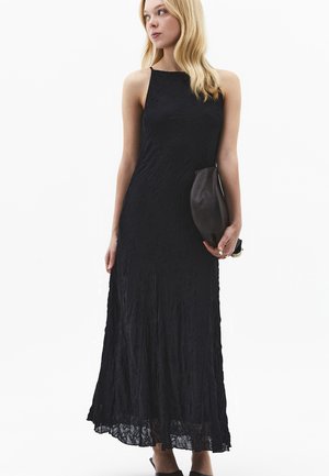 Kobieta w czarnej, teksturowanej sukni maxi bez rękawów, trzymająca czarną skórzaną kopertówkę, patrzy w lewo na białym tle.