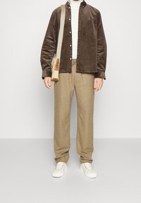 Camisa de botones de pana marrón, suéter de cuello alto blanco, pantalones de lana beige y zapatillas blancas. Bandolera con acentos verdes y marrones.