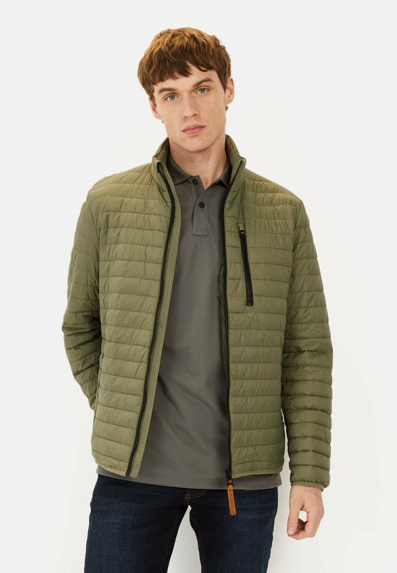 Grøn quiltet jakke med høj krave, frontlynlås og sidelommer. Stylet over en grå polo skjorte, parret med mørke jeans.