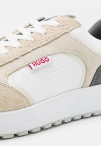 Baskets en daim beige avec des accents en mesh blanc et textile gris, dotées d'une étiquette rouge "HUGO", de lacets blancs et d'une semelle blanche texturée.