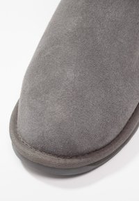 Pantofole in suede grigio con punta arrotondata e superficie texture. Presentano una suola in gomma scura a contrasto e cuciture lungo il bordo.