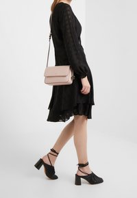 Světle růžová kožená crossbody taška s klopovým uzavíráním a zlatými detaily, kombinovaná s černými průhlednými šaty s dlouhým rukávem a černými lodičkami na kotníkovém pásku.