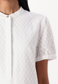 Hvit kortermet bluse med blomsterbroderi, med en mandarin-krage og en frontlås. Mykt, strukturert stoff med et delikat mønster.