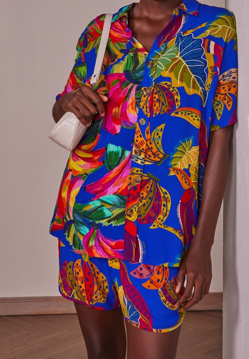 Set di camicia e pantaloni corti a motivi floreali colorati in blu, con foglie vivaci e banane, bottoni e una vestibilità rilassata. Include una piccola borsa bianca.