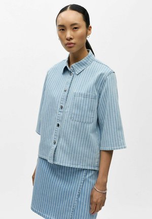 Femme aux longs cheveux noirs attachés en queue de cheval, portant une chemise à manches courtes à rayures verticales bleues et blanches, boutonnée, et une jupe assortie.