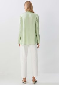 Blusa oversized verde chiaro con una texture liscia e morbida, maniche lunghe e orlo curvo, abbinata a pantaloni bianchi a gamba dritta.