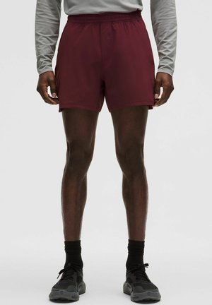 Korte, bordeauxrode sportbroekjes met een elastische tailleband, zijzakken en een gladde textuur. Draagt zwarte schoenen en sokken.