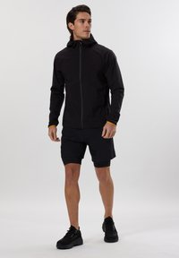 Sort letvægtsjakke med hætte, grå lynlås og orange detaljer på ærmerne, parret med sorte shorts og sorte træningssko.