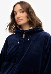 Granatowa bluza rozpinana z miękkiego materiału, z kapturem na sznurki, haftowanym logo oraz przednimi kieszeniami do przechowywania.