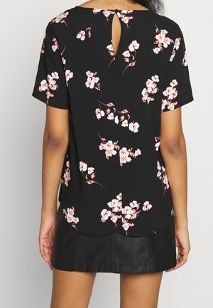 Zwart kort mouwen blouse met roze en witte bloemenprint, sleutelgatdetail aan de achterkant, gedragen met een zwarte leren rok, gezien van achteren.