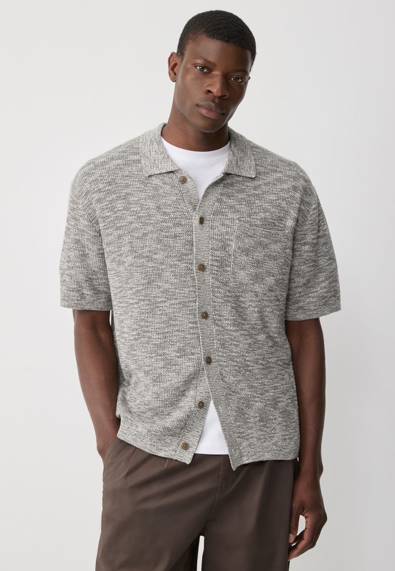 Grijs overhemd met korte mouwen en knopen, met een gestructureerde weving, voorzien van een kraag, borstzak en bruine knopen. Draagt over een wit T-shirt.