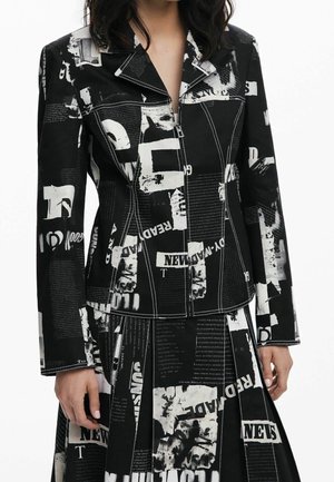 Femme portant un ensemble veste et jupe noire avec texte graphique blanc et motif imprimé journal, avec une fermeture éclair à l'avant et un col pointu.