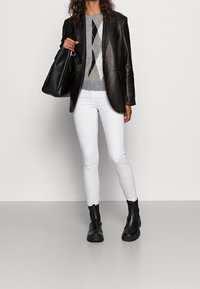 Schwarze Lederblazer über einem grauen Pullover mit Rautenmuster, weiße schmale Jeans und schwarze Ankle Boots, stehend vor einem neutralen Hintergrund.