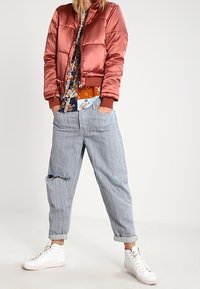 Roze satijnen bomberjack met ribbelmanchetten, bloemetjespatroon shirt, versleten lichtblauwe gestreepte jeans en witte sneakers.