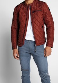 Braune gesteppte Jacke mit Stehkragen, Reißverschluss vorne und Seitentaschen. Kombiniert mit blauen, distressed Jeans mit Reißverschluss-Details.