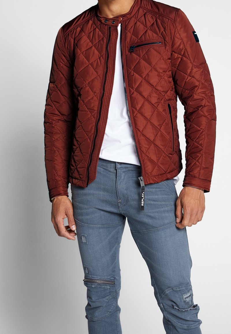 Braune gesteppte Jacke mit Stehkragen, Reißverschluss vorne und Seitentaschen. Kombiniert mit blauen, distressed Jeans mit Reißverschluss-Details.