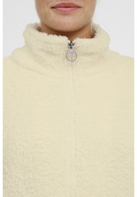 Primer plano de la parte inferior de la cara y el cuello de una persona que lleva una chaqueta con cremallera de color crema y peluda, con un tirador de cremallera metálico redondo.