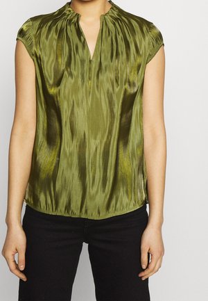 Grøn skinnende kortærmet bluse med rynket krave og slids ved halsudskæringen, båret med sorte bukser, torso og arme synlige mod hvid baggrund.