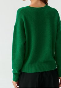 Femme portant un pull en maille vert vif et un pantalon noir, vue de dos sur un fond blanc uni.
