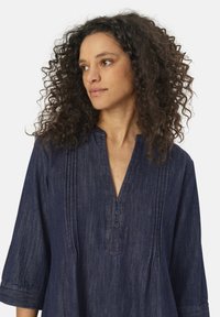 Laivastonsininen farkkutunika, jossa on V-mallinen kaula-aukko, kolme nappia ja hienovaraisia pystysuoria ompeleita. Ominaisuuksiin kuuluvat 3/4-hihat ja pehmeä rakenne.