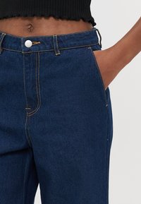 Dunkelblaue Denim-Hosen mit sichtbaren Nähten, einem Knopfverschluss vorne und Seitentaschen. Das Material ist glatt mit einem leichten Glanz.