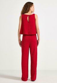 Kvinde med krøllet hår iført en ærmeløs rød glitrende top og matchende vide bukser, stående med ryggen til en ensfarvet baggrund.