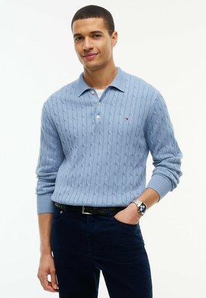 Mann trägt hellblauen Zopfpullover und dunkle Hose, mit der Hand in der Tasche, leicht lächelnd vor weißem Hintergrund.