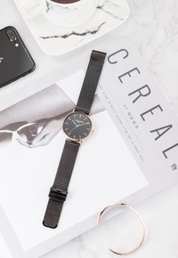 Reloj negro redondo con pulsera de malla, acentos en oro rosa, colocado sobre una revista y una superficie de mármol. Incluye un smartphone y una taza.