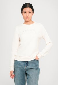 Mladá žena oblečená v krémové mikině Armani Exchange a modrých džínách, stojící s jednou rukou v kapse před jednoduchým pozadím.