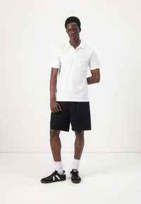 Weißes Poloshirt mit kurzen Ärmeln und einer Tasche, kombiniert mit schwarzen Shorts. Getragen mit schwarzen Schuhen und weißen Socken. Schlichtes und funktionales Design.