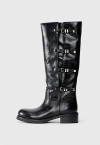 Bottines en cuir noir, hauteur genou, avec un bout arrondi, présentant trois accents de sangle avec des boucles en métal et un petit talon carré. Texture lisse.