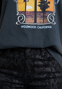 T-shirt gris foncé avec un graphisme coloré représentant un coucher de soleil et des palmiers, associé à un pantalon noir affichant un motif texturé de peau de serpent.