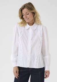 Vit blus med volangdetaljer längs knäppningen och axlarna, krage, och långa ärmar med samlade manschetter.