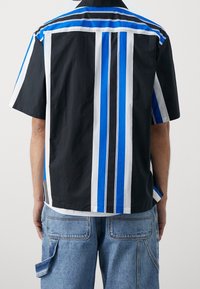 Man in een zwart shirt met korte mouwen met verticale witte en blauwe strepen, gecombineerd met lichtblauwe denim cargo shorts, met zijn rug naar ons toe.