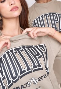 Camisetas beige con una impresión en negrita en negro que dice "MULTIPLICAR" y texto más pequeño. Cuenta con un ajuste relajado y una textura suave, mostradas en dos personas.