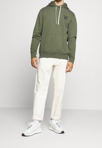 Sudadera verde oliva con bolsillo frontal, cordones y logo. Combinada con pantalones color crema, arremangados, y zapatillas blancas.