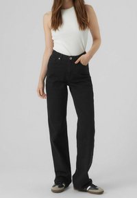 Pantalones de pierna ancha negros hechos de algodón, con una cintura alta, bolsillos frontales y un cierre clásico de botón y cremallera. Combinados con una camiseta sin mangas blanca.