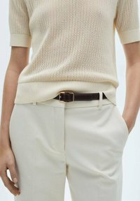 Cremefarbener Strickpullover mit kurzen Ärmeln, kombiniert mit cremefarbenen Hosen. Brauner Ledergürtel mit einer goldfarbenen Metallschnalle. Glatte Stofftextur.