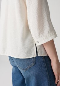 Blusa color beige chiaro, stropicciata, con maniche a tre quarti, dettaglio dei bottoni ai polsini, spacco laterale, abbinata a jeans in denim blu.