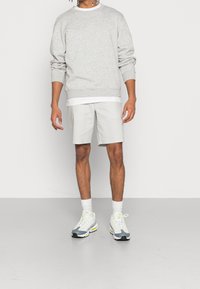 Grijze sweatshirt met een ronde hals, wit onderhemd, lichtgrijze shorts en grijze sneakers met gele accenten en witte veters.