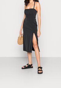 Robe midi noire en satin avec un buste froncé et de fines bretelles. Présente une fente sur le côté et une texture lisse. Portée avec des sandales noires et un sac tissé.