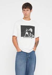 White cotton t-shirt featuring a black graphic print of two boxers on the front. Short sleeves and crew neck design. Blue jeans are worn.