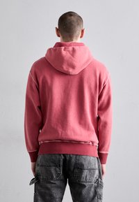 Wyblakła różowa bluza z kapturem, z kieszonką kangurką oraz ściągaczami przy rękawach i na dole, noszona z ciemnoszarymi spodniami cargo z wieloma kieszeniami.