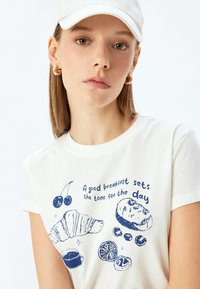 Witte katoenen T-shirt met blauwe grafische prints van voedselitems en de tekst: "Een goed ontbijt bepaalt de toon voor de dag."
