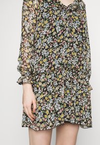 Černé květinové šaty s dlouhými nařasenými rukávy. Obsahují volánkový výstřih a živé květinové vzory v modré, růžové a žluté barvě.