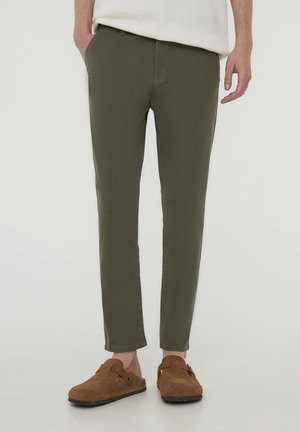 Pantaloni slim-fit verde oliva realizzati in morbido misto cotone, caratterizzati da tasche laterali e un design semplice e senza decorazioni. Indossati con scarpe beige slip-on.