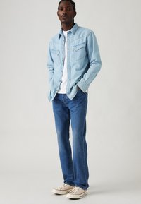 Light blue denim shirt with two chest pockets, white t-shirt underneath, blue jeans, and beige sneakers with red accents.