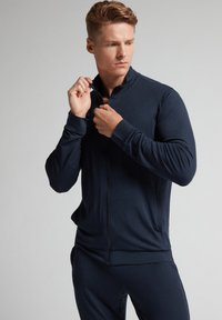 Chaqueta azul marino con cremallera hecha de tela suave, con un cuello alto, corte ajustado y mangas largas con puños acanalados.