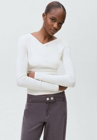 Witte longsleeve top met een V-hals en een zakje aan de linkerkant. Gepaard met grijze broek met twee knopen bij de taille.
